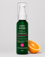 Load image into Gallery viewer, Ann Webb Vita-C Complex skincare pump bottle in dark green with a clear cap, shown next to a halved orange slice on a light gray background.