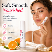 Load image into Gallery viewer, Ann Webb The Balm lip balm tube standing on a marble surface next to sliced oranges, with a woman holding the tube near her face. Text reads 'Soft, Smooth, Nourished' and lists benefits like colorless, non-greasy, and suitable for lips, face, and body.
